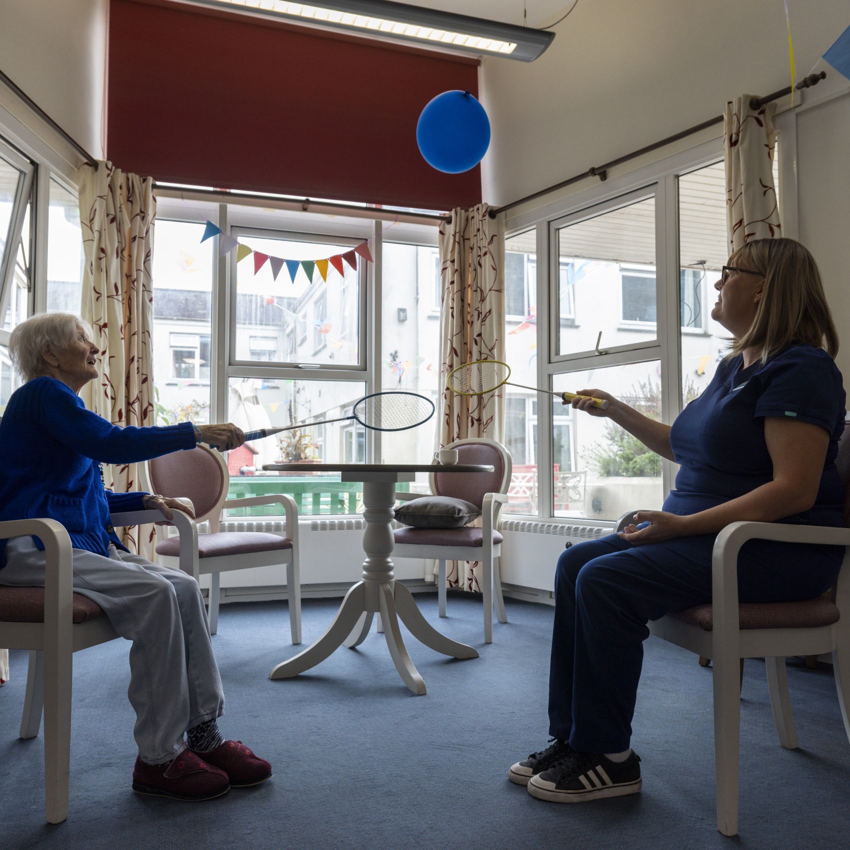 A female activity worker playing a game with badminton rackets and a balloon with an older person.