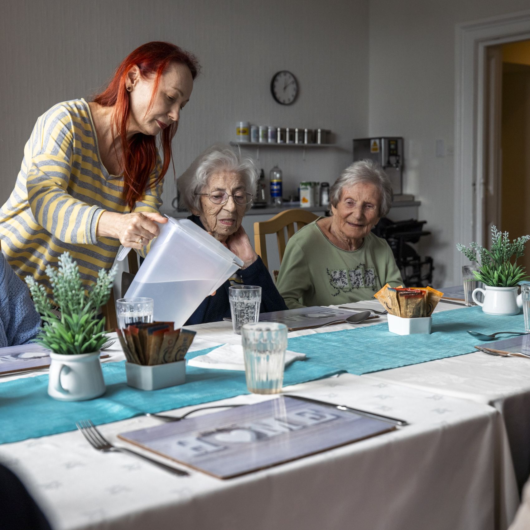 A woman pouring water from a jug into a glass at a dining table with two women sitting down