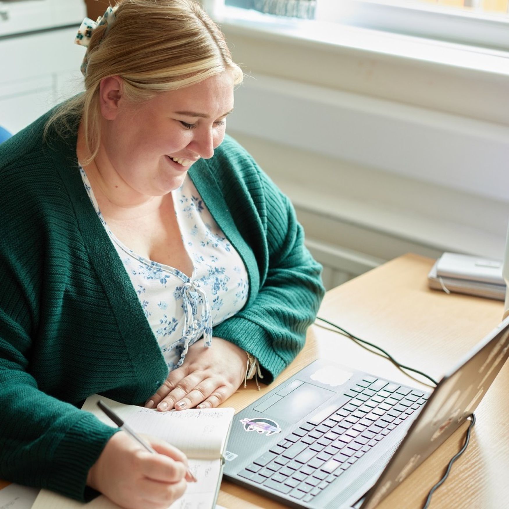 A woman writing in a notepad at a desk with a laptop