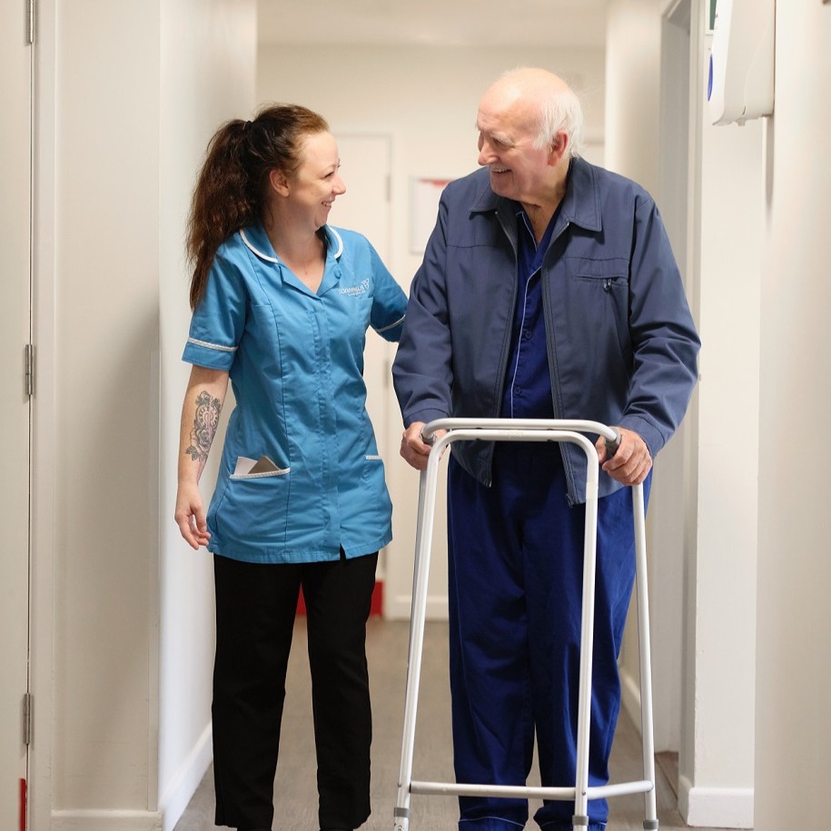 An elderly man witha walking frame helped by a female care worker