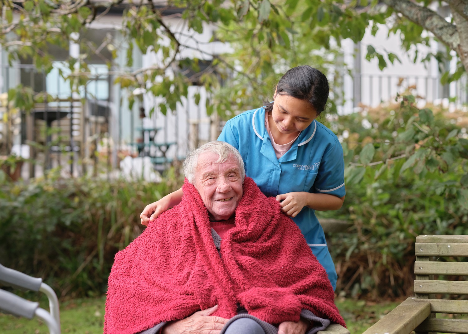 An elderly man in a wheelchair and blanket supported by a care worker
